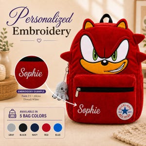 Könnte beinhalten: Ein roter Plüschrucksack mit einem Cartoon-Gesicht, großen weißen Augen, grünen Iris und einer gelben Schnauze. Der Rucksack verfügt über eine vordere Reißverschlusstasche, einen kleinen grauen Oktopus-Schlüsselanhänger und den weißen Stickschriftzug Sophie.