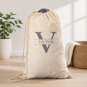 May include: A cream-colored fabric laundry bag with a drawstring closure at the top. The front features a large gray letter V with the name VICTOR printed in gray capital letters across the center of the letter, creating a personalized design.