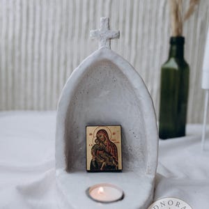 Może przedstawiać: Biały, łukowaty świecznik o wyglądzie kamienia z krzyżem na szczycie. Wewnątrz znajduje się mała, prostokątna ikona religijna przedstawiająca postać trzymającą dziecko. Zapalony podgrzewacz znajduje się w okrągłym wgłębieniu u podstawy.