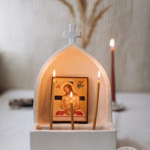 Puede incluir: Un portador de iconos de cerámica blanca con una cruz en la parte superior, que contiene un icono religioso y tres velas encendidas. Una sola vela roja arde en el fondo. El icono representa una figura religiosa. Tema religioso.