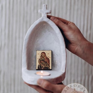 May include: A white, textured, arched ceramic candle holder featuring a cross at the top. Inside the arch, a small rectangular icon depicts a figure holding a child. A lit tea light candle sits on the base of the holder, providing a warm glow.