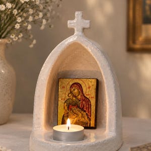 Op de afbeelding: Een wit keramieken boogvormig heiligdom met een kruis aan de bovenkant en een ingebouwde kaarsenhouder. Binnenin de boog staat een rechthoekig religieus icoon met een figuur die een kind vasthoudt, verlicht door een brandend theelichtje.