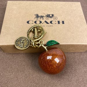 Peut inclure: Un porte-clés de la marque Coach avec une breloque en forme de pomme orange pailletée et une feuille verte, fixé à un anneau en métal doré et un médaillon circulaire gravé d'un logo de calèche, posé sur une boîte en carton beige avec le logo Coach.