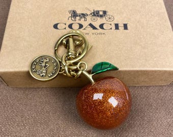 Op de afbeelding: Een Coach sleutelhanger met een ronde, glinsterende oranje appelbedel met een groen blaadje, bevestigd aan een goudkleurige metalen ring en een rond medaillon met een koetslogo, liggend op een beige kartonnen doos met het Coach-logo.