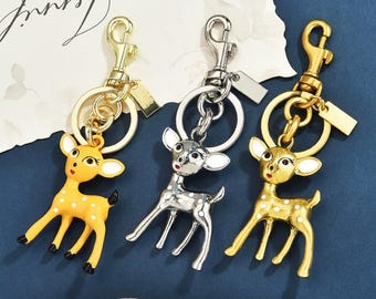 Op de afbeelding: Drie metalen sleutelhangers met hertjes met witte stippen. Eén hertje is geel, één is zilverkleurig en één is goudkleurig. Elke sleutelhanger heeft een ronde ring, een karabijnsluiting en een klein rechthoekig metalen label aan de bovenkant.