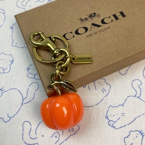 May include: A gold-tone metal keyring featuring a glossy orange pumpkin charm with a green leaf accent. The keyring includes a lobster clasp, a split ring, and a small rectangular tag engraved with the word COACH. It rests on a brown box.