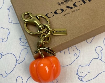 Op de afbeelding: Een goudkleurige metalen sleutelhanger met een glanzende oranje pompoenbedel en een groen blaadje. De sleutelhanger bevat een ronde ring, een karabijnsluiting en een klein rechthoekig label met de tekst COACH NEW YORK.