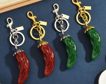 Op de afbeelding: Vier sleutelhangers met chilipeperbedels. Twee sleutelhangers hebben zilverkleurige hardware en twee hebben goudkleurige hardware. De chilipepers zijn glanzend, twee in rood en twee in groen, elk met een gestructureerde metalen dop.