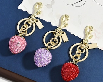 Op de afbeelding: Drie aardbeivormige sleutelhangers met goudkleurige metalen onderdelen, waaronder een karabijnhaak, een ronde ring en een klein rechthoekig label. Elke aardbei-bedel is bedekt met kleine, sprankelende kristallen in roze, paars en rood.