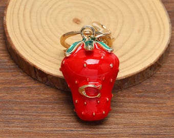 Op de afbeelding: Een felrode aardbeivormige bedel met een goudkleurige metalen afwerking en groene emaille blaadjes aan de bovenkant. De aardbei is voorzien van een kleine gouden letter C op de voorkant en een goudkleurige metalen ring aan de bovenkant.