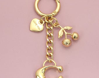 Op de afbeelding: Een goudkleurige metalen sleutelhanger met een ronde ring, een ketting, een hartvormige bedel met de tekst LOVE YOU, een kersenbedel met twee getextureerde vruchten en blaadjes, en een grote letter C aan de onderkant.