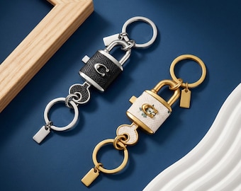 Op de afbeelding: Twee hangslotvormige sleutelhangers met zilver- en goudkleurige hardware. De een heeft zwart gestructureerd leer met een zilveren C-logo, de ander wit leer met bloemenpatroon en een gouden C-logo. Beide bevatten een sleutel, een ring en een label.