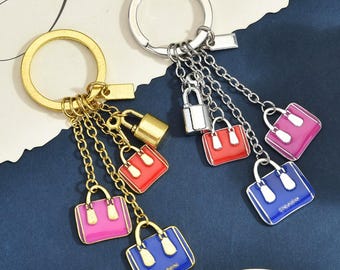 Op de afbeelding: Twee metalen sleutelhangers, één goudkleurig en één zilverkleurig, elk met een ronde ring bevestigd aan meerdere kettinkjes. Elke ketting heeft een klein hangslotje en twee miniatuur handtasbedels in felrood, roze en blauw emaille.