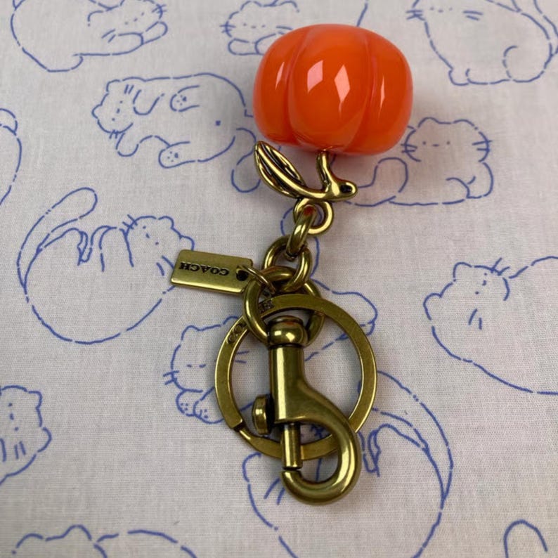 May include: A bright orange pumpkin-shaped charm attached to a brass-toned metal keychain. The keychain features a circular ring, a lobster claw clasp, and a small rectangular metal tag engraved with the word COACH in capital letters.
