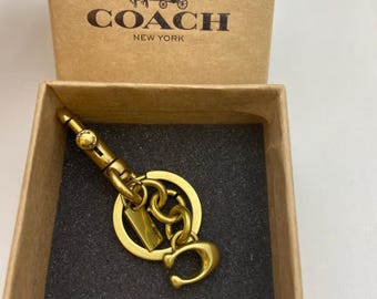 Op de afbeelding: Een goudkleurige metalen sleutelhanger met een karabijnhaak, een ronde ring en een C-vormige bedel, gepresenteerd in een beige kartonnen doos met het Coach New York logo en een koetsafbeelding gedrukt op de binnenkant van het deksel.