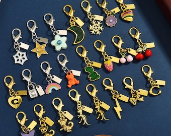 Op de afbeelding: Een verzameling metalen bedels met karabijnsluiting in diverse emaille en metalen vormen, waaronder een maan, sneeuwvlok, bloem, hart, ster, regenboog, dinosaurus, beer, kat, vlinder, bliksemschicht, strik en kers, elk voorzien van een klein leeg label.