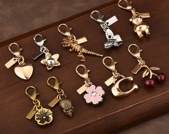 Op de afbeelding: Een verzameling van tien metalen bedels met karabijnsluitingen, waaronder vormen zoals een hart, een ster, een dinosaurusskelet, een vlinder, een teddybeer, een bloem, een muis, een roze klavertjevier, een letter C en een paar rode kersen.