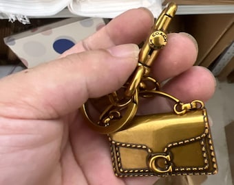 May include: A metallic gold-toned keychain featuring a miniature handbag charm with a structured flap, a prominent letter C logo on the front, and decorative stitching details. The charm is attached to a circular ring and a swivel lobster clasp.