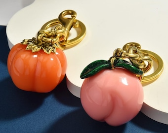 Op de afbeelding: Twee sleutelhangers met fruitvormige bedels aan goudkleurige metalen ringen en sluitingen. De ene bedel is een glanzende oranje pompoen met een gouden blad, de andere een gladde lichtroze perzik met donkergroene geëmailleerde blaadjes.