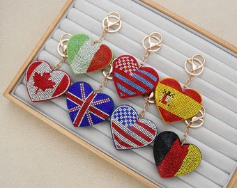 Op de afbeelding: Zes hartvormige sleutelhangers versierd met kleurrijke strass-steentjes, elk met een ander nationaal vlagontwerp, waaronder Canada, Italië, het Verenigd Koninkrijk, de VS, Spanje en Duitsland, bevestigd aan goudkleurige metalen ringen.