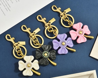 Op de afbeelding: Vier metalen sleutelhangers met goudkleurige karabijnsluitingen, ringen en kleine rechthoekige labels. Elke sleutelhanger heeft een klavertjevier in wit, zwart glitter, lavendel of roze, met een centraal cluster van kleine kristallen.