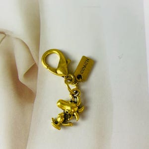 Puede incluir: Un colgante de metal dorado para bolso con cierre de mosquetón y una etiqueta rectangular grabada con la palabra COACH. Unido al cierre hay un pequeño dije detallado que representa a un personaje de perro de dibujos animados.