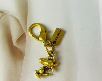 Op de afbeelding: Een goudkleurige metalen tashanger met een karabijnsluiting en een rechthoekig label waarop het woord COACH is gegraveerd. Aan de sluiting hangt een kleine, gedetailleerde bedel in de vorm van een stripfiguurhond in een staande houding.