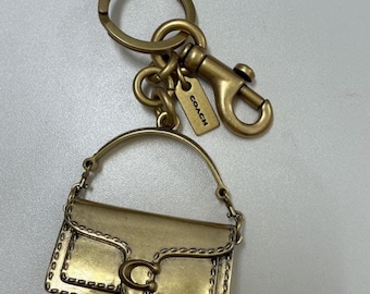 May include: A brass-toned metal keyring featuring a miniature handbag charm with a flap closure and a prominent C-shaped logo. The keyring includes a circular ring, a lobster clasp, and a small rectangular tag, each engraved with the word COACH.