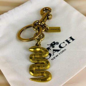 May include: A gold-toned metal keyring featuring a coiled snake charm with textured scales. The keyring includes a circular ring, a lobster claw clasp, and a small rectangular tag engraved with the word COACH. The metal hardware has a polished finish.
