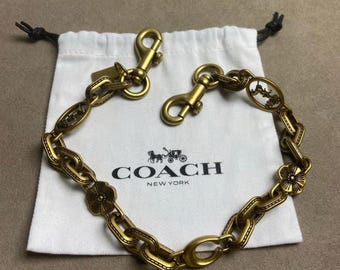 Op de afbeelding: Een goudkleurige metalen ketting met ovale schakels, bloemvormige bedels en twee karabijnsluitingen. De ketting ligt op een wit stoffen trekkoordzakje bedrukt met het zwarte Coach New York logo en een icoon van een paard en wagen.