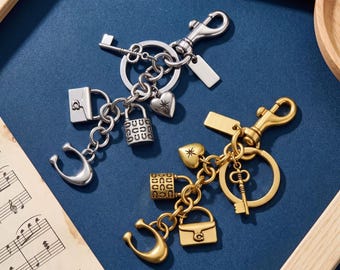Op de afbeelding: Twee metalen sleutelhangers, één zilverkleurig en één goudkleurig, elk voorzien van een karabijnhaak, een ring en diverse bedels waaronder een kleine handtas, een hangslot, een sleutel, een hartje en een C-vormig logo op blauw.