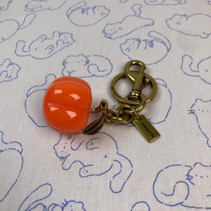 May include: A bright orange pumpkin-shaped charm attached to a gold-tone metal keychain. The keychain features a sturdy lobster clasp, a circular ring, and a small rectangular metal tag engraved with the word COACH in capital letters.