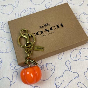 May include: A brass-toned metal keychain featuring a glossy orange pumpkin charm and a small rectangular tag embossed with the word COACH. The keychain is attached to a lobster clasp and rests on a brown cardboard box printed with the COACH NEW YORK logo.