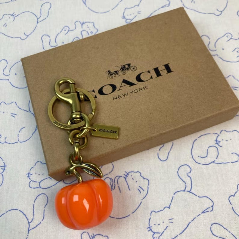 May include: A brass-toned metal keychain featuring a glossy orange pumpkin charm and a small rectangular tag embossed with the word COACH. The keychain is attached to a lobster clasp and rests on a brown cardboard box printed with the COACH NEW YORK logo.