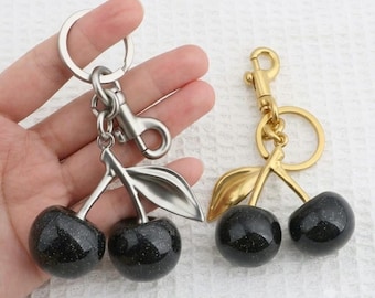 Op de afbeelding: Twee kersvormige sleutelhangers, één met een zilverkleurige metalen afwerking en één met een goudkleurige metalen afwerking. Elke sleutelhanger heeft twee ronde, donkere, glinsterende bollen die kersen voorstellen, bevestigd aan een metalen steeltje.