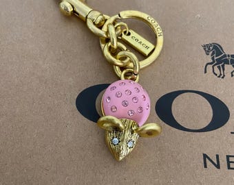 Op de afbeelding: Een goudkleurige sleutelhanger met een muisbedel, voorzien van een roze kop met kristallen en een gestructureerd goudkleurig lichaam met kristallen ogen. De sleutelhanger bevat een karabijnhaak, een ring en een label met de inscriptie COACH.