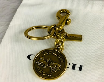 Op de afbeelding: Een goudkleurige metalen sleutelhanger met een ronde bedel voorzien van een paard-en-wagenmotief en het woord COACH. De sleutelhanger bevat een splitring, een karabijnsluiting en een klein rechthoekig label met de merknaam COACH.