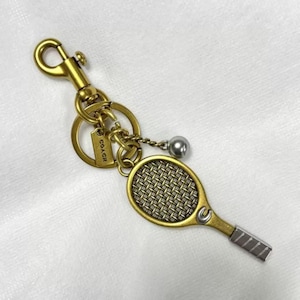 May include: A gold-toned metal keyring featuring a tennis racket charm with a textured mesh head, a small silver-toned ball charm, and a circular ring attached to a lobster clasp. A rectangular tag engraved with the word COACH is attached to the ring.