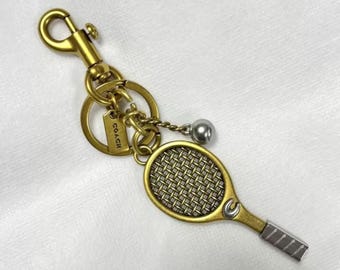 May include: A gold-toned metal keychain featuring a tennis racket charm with a textured mesh head, a small silver-toned ball charm, and a circular ring attached to a lobster clasp. A rectangular tag engraved with the word COACH is attached to the ring.
