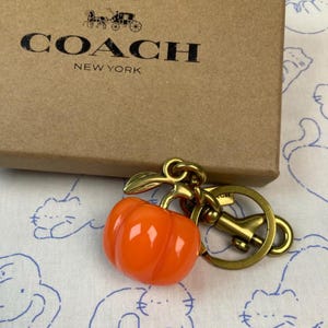 May include: A bright orange pumpkin-shaped charm attached to a brass-toned metal keyring and lobster clasp. The charm is positioned in front of a tan cardboard box featuring the black Coach New York logo and a horse and carriage graphic.