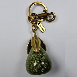 May include: A pear-shaped keyring featuring a glittery olive green body with a metallic brass-toned stem and leaf. The hardware includes a circular ring and a lobster clasp, with a small rectangular tag engraved with the word COACH attached.