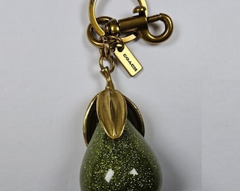 May include: A pear-shaped keychain featuring a glittery olive green body with a metallic brass-toned stem and leaf. The hardware includes a circular ring and a lobster clasp, with a small rectangular tag engraved with the word COACH attached.