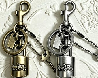 Op de afbeelding: Twee metalen sleutelhangers met een hangslotontwerp en een paard-en-wagen embleem. De ene is afgewerkt in antiek messing, de andere in gepolijst zilver. Elke hanger bevat een karabijnhaak, een sleutelring en een klein sleuteltje aan een kettinkje.
