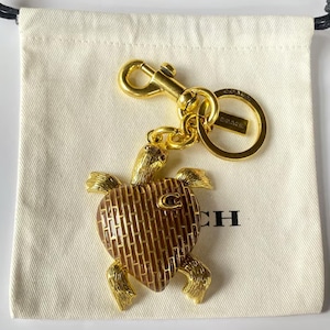 May include: A gold-tone metal turtle keyring featuring a heart-shaped shell with a brown brick-like pattern and a small letter C emblem. The turtle has textured limbs and head, attached to a gold-tone lobster clasp and a round split ring with Coach branding.