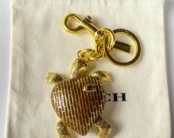 Op de afbeelding: Een goudkleurige schildpad sleutelhanger met een hartvormig schild voorzien van een bruin baksteenpatroon en een klein letter C-embleem. De schildpad heeft gestructureerde ledematen en een kop, bevestigd aan een goudkleurige karabijnhaak en ring met Coach-merk.