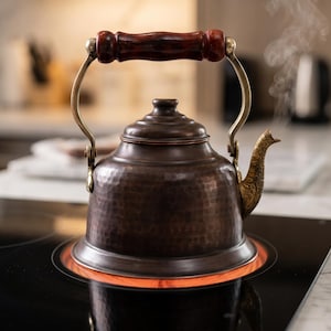 May include: A copper kettle with a dark wooden handle and brass accents sits on a black stovetop. Steam rises from the spout, indicating the water is hot. The kettle has a hammered texture and a decorative spout.
