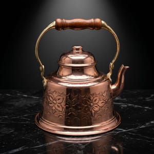 Puede incluir: Una tetera de cobre con un diseño floral, detalles de latón y un asa de madera. La tetera se asienta sobre una base redonda y tiene un pico para verter. El asa es arqueada y se sujeta con accesorios de latón.