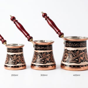 May include: Three copper coffee pots with dark wood handles and floral patterns. The pots are labelled with their capacity: 200ml, 300ml, and 400ml. The pots have a silver rim and a copper body with black and white accents.