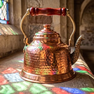 May include: A hammered copper tea kettle featuring a polished finish, a rounded body, and a lid with a stepped design. The kettle has a curved spout with embossed floral detailing and a brass-toned handle with a dark wooden grip at the top.