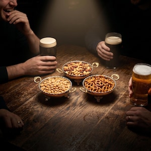 Può includere: Un tavolo di legno con tre ciotole decorative piene di snack: arachidi, frutta secca mista e pretzel. Diverse persone tengono bicchieri di birra, con una persona che ride. La scena è debolmente illuminata.